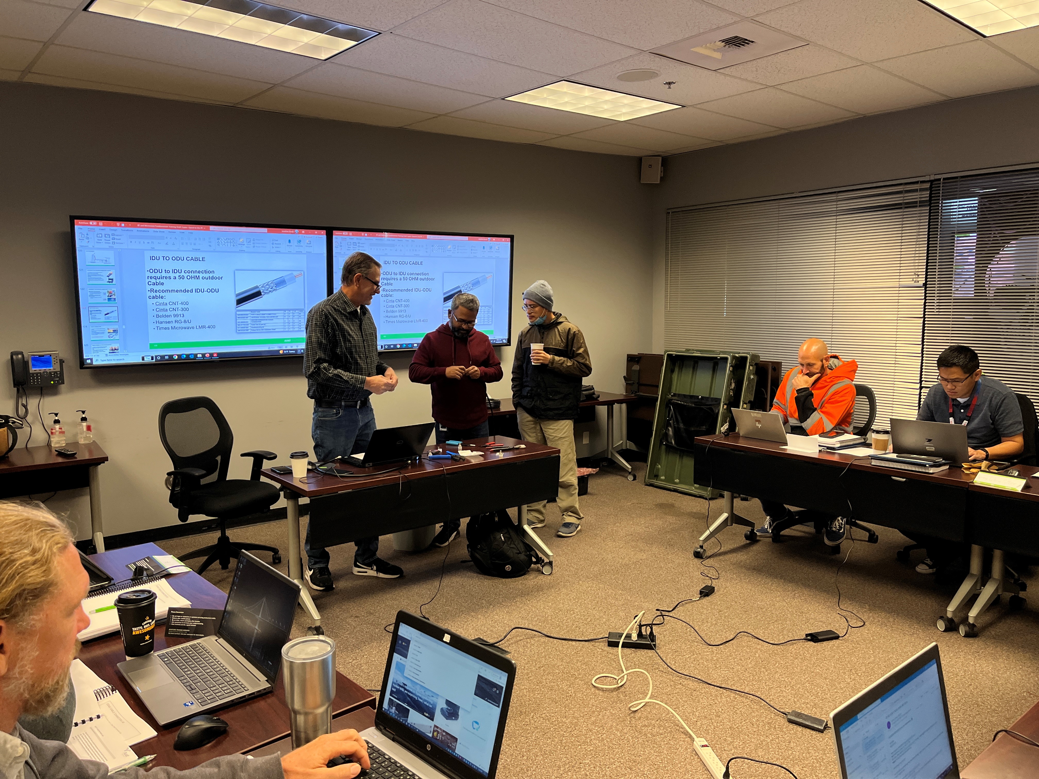 Caltrans ITS engineers practice cable and connector preparation as a lab exercise during the RF Fundamentals training. Three people stand in the front of a classroom. One person is holding cables as part of a demonstration.